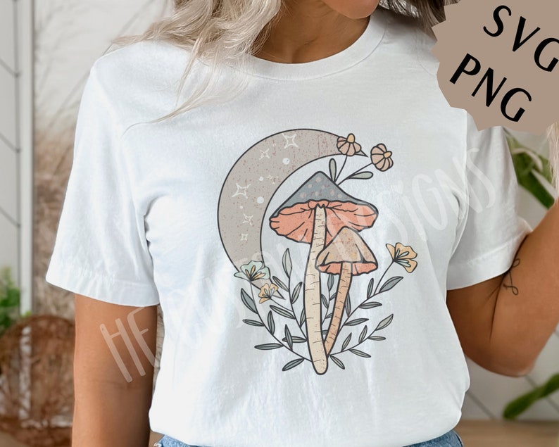 May include: White t-shirt with a graphic of two mushrooms, a crescent moon, and flowers. The mushrooms are orange and brown with white spots. The moon is gray with white stars. The flowers are pink, yellow, and white.