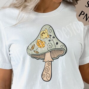 May include: A white t-shirt with a green and yellow mushroom graphic. The mushroom has a swirl design and is decorated with flowers.