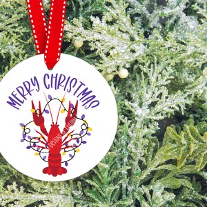 May include: White round Christmas ornament with a red crawfish holding purple and yellow Christmas lights. The ornament reads "Merry Christmas!" in purple text.