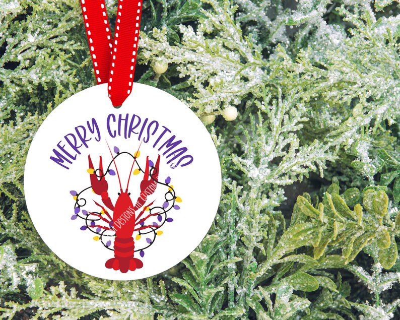 May include: White round Christmas ornament with a red crawfish holding purple and yellow Christmas lights. The ornament reads "Merry Christmas!" in purple text.
