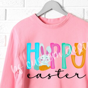 May include: Pink sweatshirt with the text "Happy Easter" in colorful letters. A cartoon bunny holding a carrot is in the middle of the word "Happy".