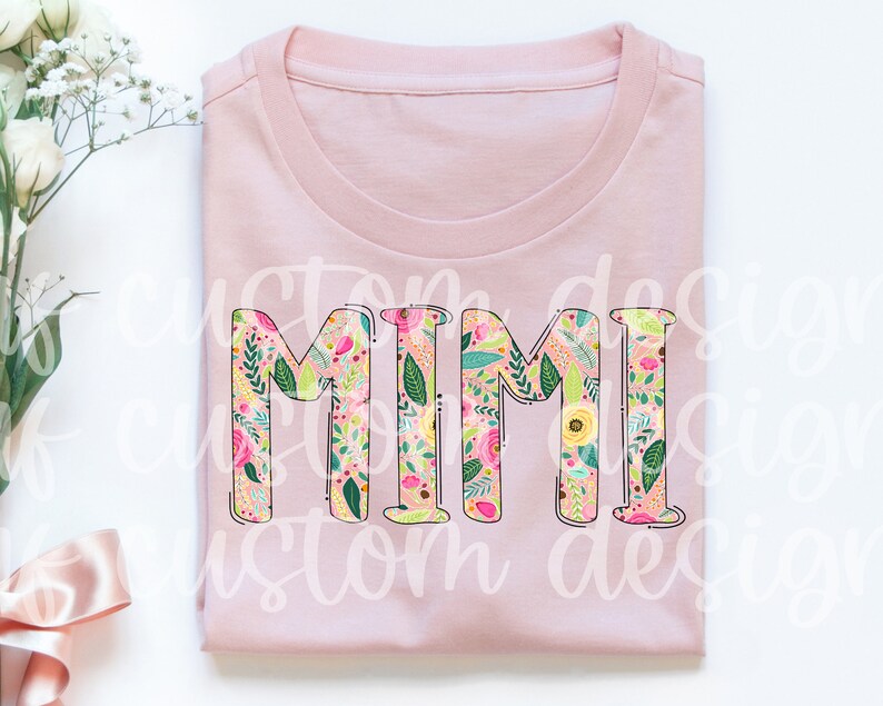 Pu&ograve; includere: Maglietta rosa con la parola "MIMI" in grandi lettere contornate. Le lettere sono riempite con un motivo floreale in tonalit&agrave; rosa, giallo, verde e bianco.