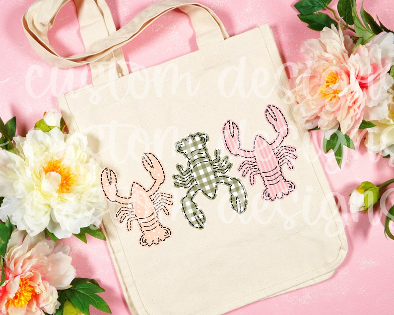 May include: A white canvas tote bag with three embroidered lobsters. The lobsters are in different colors and patterns: pink with white stripes, green with white gingham, and pink with white polka dots.