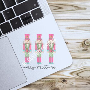 May include: Three pink nutcracker figurines with green and white accents stand in a row on a white background. The text "merry christmas" is written below the figurines.