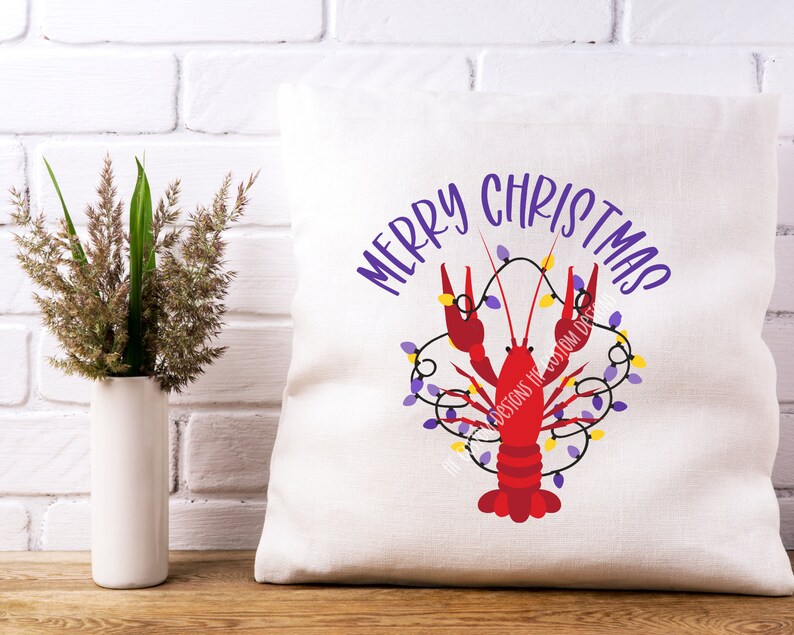 May include: A white pillow with the text "Merry Christmas" in purple lettering. A red crawfish is decorated with purple and yellow lights.