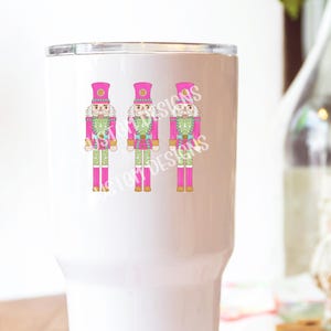 K&ouml;nnte beinhalten: Ein wei&szlig;er Becher mit drei pinkfarbenen und gr&uuml;nen Nussknacker-Illustrationen. Die Nussknacker tragen rosa H&uuml;te und haben goldene Akzente.