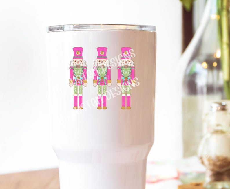 K&ouml;nnte beinhalten: Ein wei&szlig;er Becher mit drei pinkfarbenen und gr&uuml;nen Nussknacker-Illustrationen. Die Nussknacker tragen rosa H&uuml;te und haben goldene Akzente.