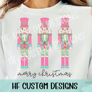May include: Three pink and white nutcracker figurines with green and blue accents. The nutcrackers are standing in a row and have the text "merry christmas" below them. The text "HF CUSTOM DESIGNS" is at the bottom of the image.