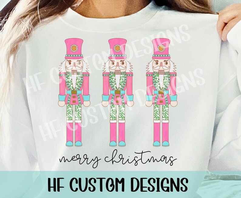 May include: Three pink and white nutcracker figurines with green and blue accents. The nutcrackers are standing in a row and have the text "merry christmas" below them. The text "HF CUSTOM DESIGNS" is at the bottom of the image.