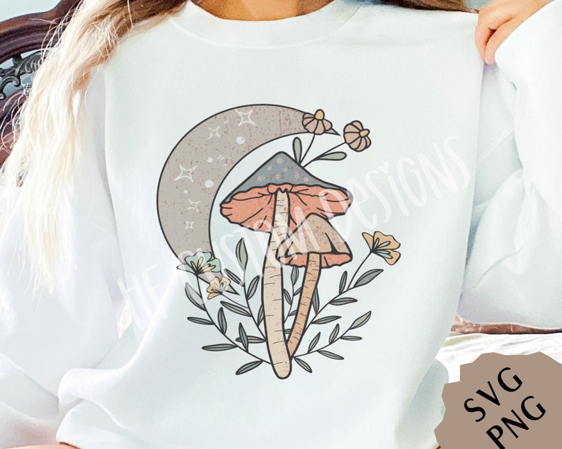 May include: A white sweatshirt with a graphic design of two mushrooms with a crescent moon and flowers. The mushrooms are pink and brown with white spots. The flowers are pink, yellow, and blue.