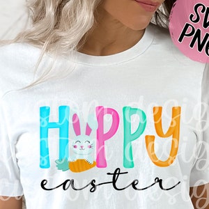 May include: White t-shirt with the text "Happy Easter" in colorful letters. A cartoon bunny holding a carrot is in the middle of the word "Happy".