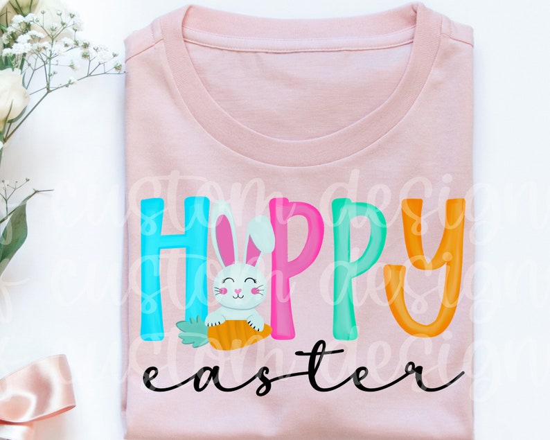 May include: A pink t-shirt with the text "Happy Easter" in colorful letters. A white bunny holding a carrot is in the middle of the word "Happy".