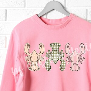 May include: Pink sweatshirt with three embroidered lobster designs. The lobsters are in different colors and patterns, including pink, green, and white.