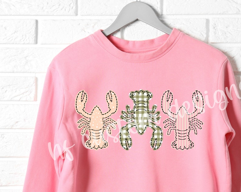 May include: Pink sweatshirt with three embroidered lobster designs. The lobsters are in different colors and patterns, including pink, green, and white.