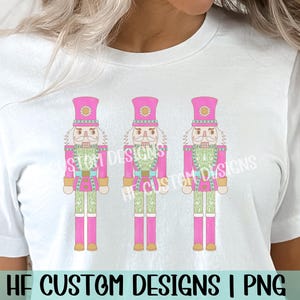 K&ouml;nnte beinhalten: Ein wei&szlig;es T-Shirt mit einem Design aus drei rosa und gr&uuml;nen Nussknacker-Soldaten. Die Nussknacker tragen rosa H&uuml;te und haben wei&szlig;e B&auml;rte. Der Text "HF CUSTOM DESIGNS | PNG" befindet sich am unteren Rand des Shirts.
