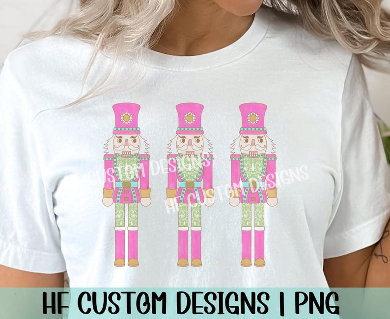 K&ouml;nnte beinhalten: Ein wei&szlig;es T-Shirt mit einem Design aus drei rosa und gr&uuml;nen Nussknacker-Soldaten. Die Nussknacker tragen rosa H&uuml;te und haben wei&szlig;e B&auml;rte. Der Text "HF CUSTOM DESIGNS | PNG" befindet sich am unteren Rand des Shirts.