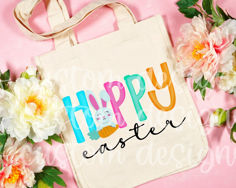 May include: A white canvas tote bag with a colorful "Happy Easter" design featuring a cartoon bunny holding a carrot. The letters are in different colors and the word "Easter" is in a cursive font.