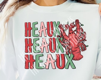May include: White sweatshirt with the text "Heaux" repeated three times in green, pink, and red lettering. A red lobster with green holly is centered on the shirt.