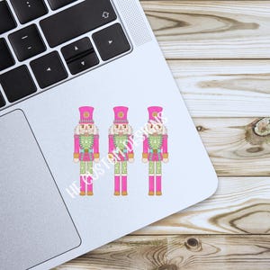 K&ouml;nnte beinhalten: Drei rosa und gr&uuml;ne Nussknackerfiguren mit goldenen Akzenten. Die Nussknacker stehen in einer Reihe auf wei&szlig;em Hintergrund.
