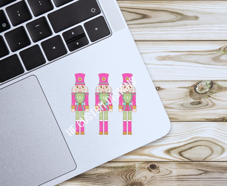 K&ouml;nnte beinhalten: Drei rosa und gr&uuml;ne Nussknackerfiguren mit goldenen Akzenten. Die Nussknacker stehen in einer Reihe auf wei&szlig;em Hintergrund.