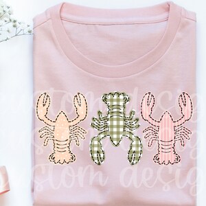 May include: A pink t-shirt with three embroidered lobsters. The lobsters are in different patterns: one is pink and white stripes, one is green and white gingham, and one is peach with white polka dots.