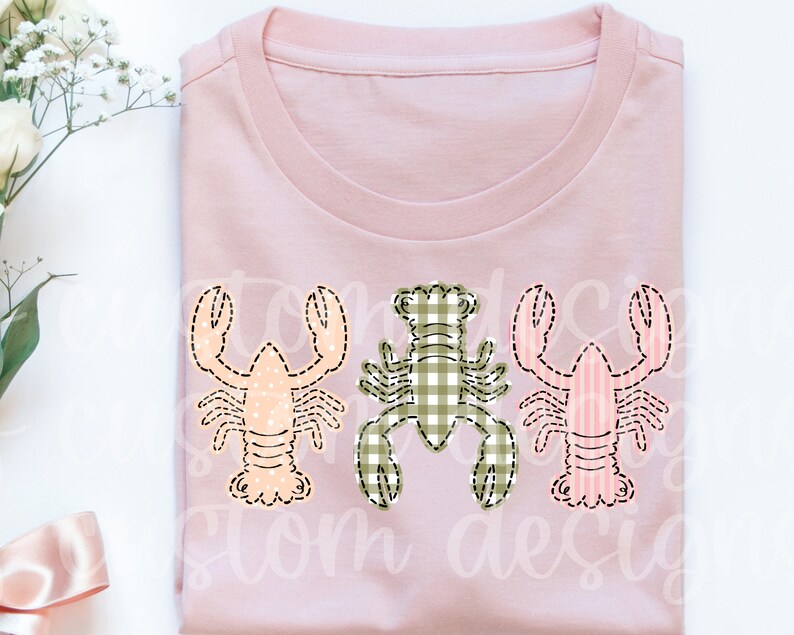 May include: A pink t-shirt with three embroidered lobsters. The lobsters are in different patterns: one is pink and white stripes, one is green and white gingham, and one is peach with white polka dots.