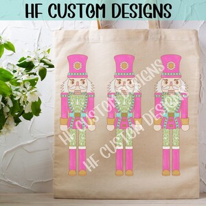 K&ouml;nnte beinhalten: Drei rosa und gr&uuml;ne Nussknackerfiguren mit wei&szlig;en Gesichtern und schwarzen Augen. Die Nussknacker stehen auf einer beigefarbenen Tragetasche mit dem Text "HF CUSTOM DESIGNS" obenauf.