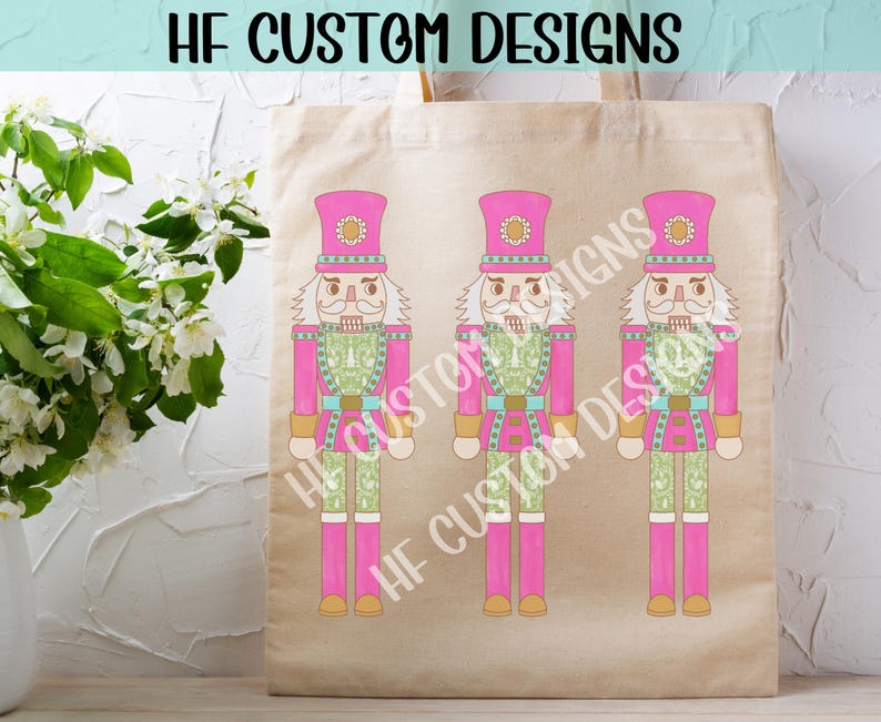 K&ouml;nnte beinhalten: Drei rosa und gr&uuml;ne Nussknackerfiguren mit wei&szlig;en Gesichtern und schwarzen Augen. Die Nussknacker stehen auf einer beigefarbenen Tragetasche mit dem Text "HF CUSTOM DESIGNS" obenauf.