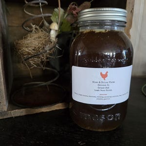 May include: A glass Mason jar filled with a dark brown liquid, labeled "Rise & Shine Farm Lamb Bone Broth." The jar has a silver lid and sits on a dark surface. Decorative elements include a wooden box and a metal spring.