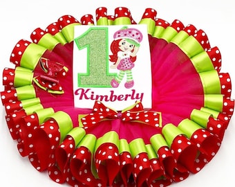 May include: A pink tulle tutu with red and green ribbon trim. The tutu has white polka dots on the red ribbon. A white onesie with a green number one and a strawberry character is on top of the tutu. The onesie also has the name Kimberly on it.
