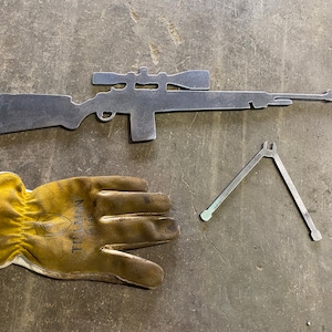 Op de afbeelding: Een metalen silhouet van een geweer met een richtkijker, een tang van metaal en een gele werkhandschoen met de tekst "Tillman" erop gedrukt.