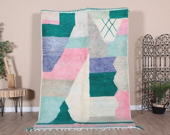 May include: A white, hand-woven rug with geometric patterns in shades of pink, blue, green, and grey. The rug has a fringe border.
