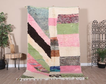 May include: A large, colourful rug with a geometric design. The rug features a mix of pink, green, black, and white stripes and shapes. The rug has a textured appearance and is displayed against a neutral wall.