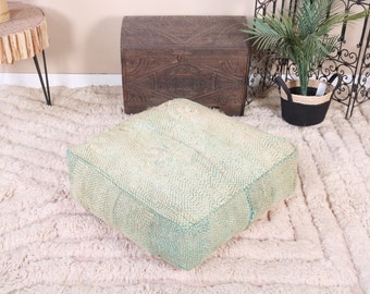 May include: A light green square floor cushion with a textured surface. The cushion is sitting on a white fluffy rug.