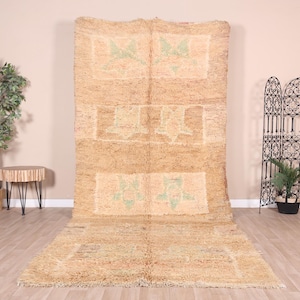 May include: A large, rectangular, beige rug with a woven pattern of green and white floral designs. The rug has a distressed, vintage look and is made of natural fibers.