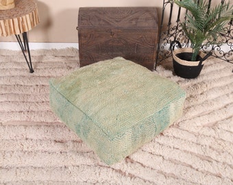 May include: A light green square floor cushion with a textured woven fabric. The cushion is sitting on a white fluffy rug.