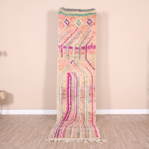 May include: A long, rectangular rug with a cream-colored base and colorful geometric patterns. The design includes pink, purple, and blue vertical stripes and diamond shapes. The rug has a fringed edge and is suitable for a hallway or entryway.