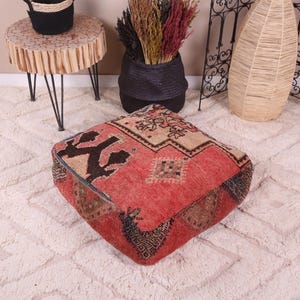 May include: A square floor pouf featuring a vibrant red base with intricate geometric patterns in black, beige, and brown. The textile surface displays a woven texture with symmetrical motifs and a dark border, providing a low-profile seat.