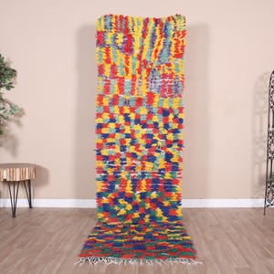 May include: A colorful, rectangular rug with a vibrant patchwork design. The rug features a mix of red, yellow, blue, and green hues. The rug is made of a textured material and has a fringe at the bottom. The rug is displayed on a wooden floor.