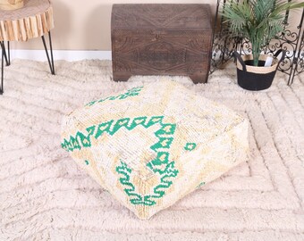 May include: A square, cream-colored pouf with a geometric green design. The pouf is made of a textured fabric and sits on a cream-colored rug. The pouf is a decorative piece of furniture.