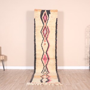 May include: A long, narrow, cream-colored rug with a black, pink, and orange geometric pattern. The rug has fringe on the ends.
