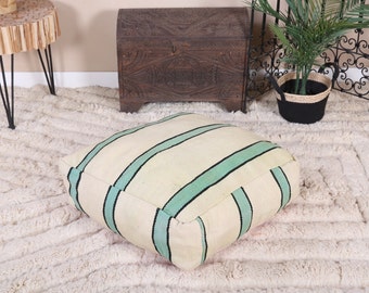 May include: Square floor cushion with a cream-colored base and vertical stripes in light teal and black. The cushion is made of a woven fabric and is placed on a textured, off-white rug. The cushion is a decorative element for a living room or bedroom.