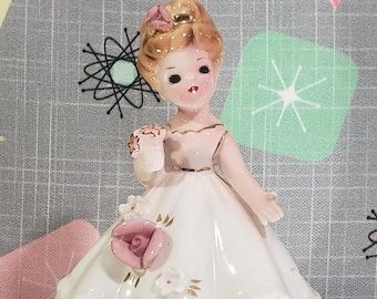 Pode incluir: Uma figura de porcelana de uma menina vestindo um vestido branco com detalhes dourados e uma rosa rosa. Ela tem cabelo loiro e uma flor rosa no cabelo.