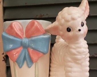 May include: A white ceramic lamb figurine with blue and pink accents. The lamb is holding a planter with a blue and pink bow. The planter has a white base with green stripes.