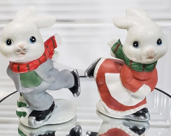 May include: Two white porcelain bunny figurines ice skating. The bunny on the left is wearing a red and green scarf and gray pants. The bunny on the right is wearing a red and green scarf and a red dress. Both bunnies have black ice skates.