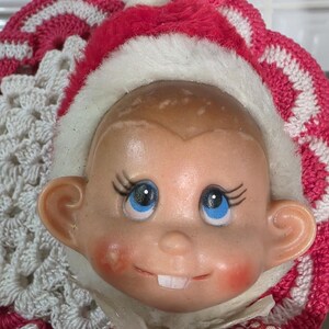 May include: A vintage doll head with blue eyes, rosy cheeks, and two front teeth. The doll wears a pink and white crocheted hat with a white fur trim and a red and white polka dot dress.