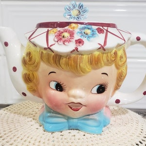 May include: A white ceramic teapot shaped like a girl's head with a blue bow, wearing a white hat with pink and blue flowers. The teapot has red polka dots on the spout and handle.