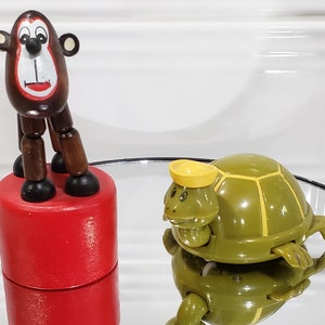 May include: A wooden monkey toy with a red face and brown body, standing on a red base. A green and yellow wind-up turtle toy is on a glass surface.
