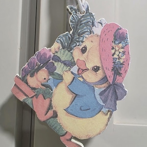 May include: A vintage-style Easter ornament featuring a yellow duckling wearing a blue jacket and a pink bonnet adorned with flowers. The duckling is holding a green gift and is surrounded by floral and foliage elements. A white string is attached for hanging.