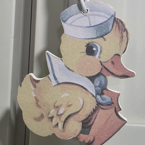May include: A yellow duck-shaped ornament wearing a sailor hat and uniform. The duck has a light blue hat, a red-orange beak, and a blue bow tie. The ornament is hanging from a white string.